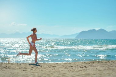 Side view of active sport woman running in morning along the beach. Fit girl jogging on seashore with mountains, cardio workout training outdoors