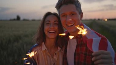 close up sparklers portrait of happy caucasian couple woman and man celebrating enjoying independence day celebration in field at sunset. couple wrapping in USA national flag, 4th of july