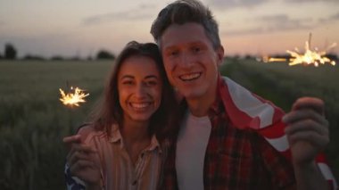 close up sparklers portrait of happy caucasian couple woman and man celebrating enjoying independence day celebration in field at sunset. couple wrapping in USA national flag, 4th of july
