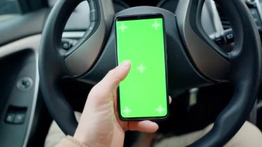 POV man in car at driver place, holds a phone with green screen with marks in hand. driver holds phone against steering wheel, swiping by finger in different directions. navigation, shopping concept