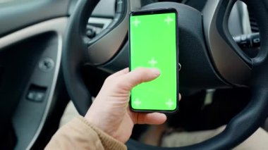 POV man in car at driver place, holds a phone with green screen with marks in hand. driver holds phone against steering wheel, swiping by finger in different directions. navigation, shopping concept