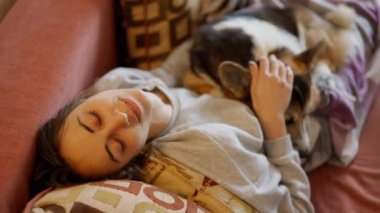 relaxed young woman with lazy dog welsh corgi breed lying on her, lies together on couch in living room. siesta, rest time at home with lovely pet