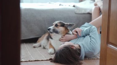 woman lying on floor in bedroom with her cute funny welsh corgi dog and stroking him. friendship with pets, games together and leisure. domestic animals in peoples life
