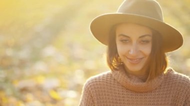close up autumn portrait of beautiful smiling woman wearing in wrown dress, sweater and hat. girl holds yellow leaves and playfuly looking at camera