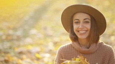 close up autumn portrait of beautiful smiling woman wearing in wrown dress, sweater and hat. girl holds yellow leaves and playfuly looking at camera