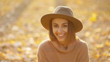 close up autumn portrait of beautiful smiling woman wearing in wrown dress, sweater and hat. girl holds yellow leaves and playfuly looking at camera