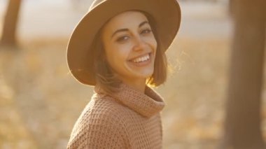 close up autumn portrait of beautiful smiling woman wearing in wrown dress, sweater and hat. girl holds yellow leaves and playfuly looking at camera