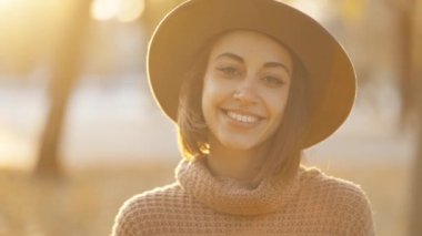 close up autumn portrait of beautiful smiling woman wearing in wrown dress, sweater and hat. girl holds yellow leaves and playfuly looking at camera