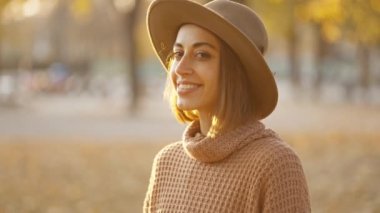 close up autumn portrait of beautiful smiling woman wearing in wrown dress, sweater and hat. girl holds yellow leaves and playfuly looking at camera