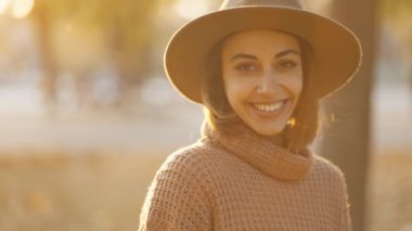 close up autumn portrait of beautiful smiling woman wearing in wrown dress, sweater and hat. girl holds yellow leaves and playfuly looking at camera