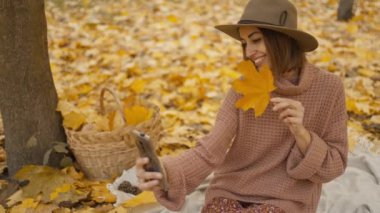 pretty young brunette woman in hat sits in autumn park with yellow foliage. joyful female makes selfie with maple leaf, or filming video, call. attractive girl in park at warm fall day. slow motion