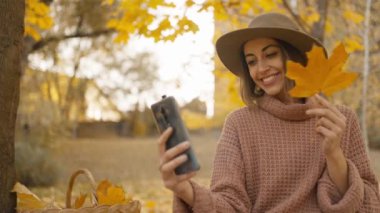 pretty young brunette woman in hat sits in autumn park with yellow foliage. joyful female makes selfie with maple leaf, or filming video, call. attractive girl in park at warm fall day. slow motion