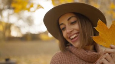 pretty young brunette woman in hat sits in autumn park with yellow foliage. joyful female makes selfie with maple leaf, or filming video, call. attractive girl in park at warm fall day. slow motion