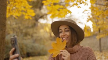 pretty young brunette woman in hat sits in autumn park with yellow foliage. joyful female makes selfie with maple leaf, or filming video, call. attractive girl in park at warm fall day. slow motion