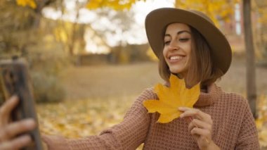 pretty young brunette woman in hat sits in autumn park with yellow foliage. joyful female makes selfie with maple leaf, or filming video, call. attractive girl in park at warm fall day. slow motion