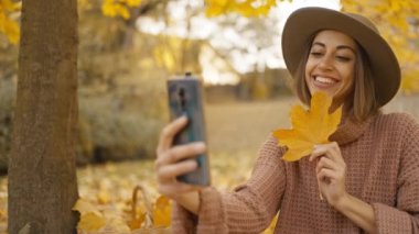 pretty young brunette woman in hat sits in autumn park with yellow foliage. joyful female makes selfie with maple leaf, or filming video, call. attractive girl in park at warm fall day. slow motion