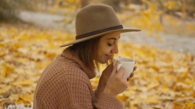 close up side view of charming happy brunette woman at picnic in bright autumn park with yellow foliage. stylish attractive girl sits on blanket in park at warm fall day and drinks coffee.