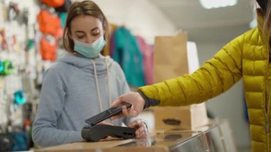 Tezgâhtaki müşteri kadın satış asistanından kıyafet satın alıyor, Temassız NFC Akıllı Telefon Dokunma Terminali ile ödeme yapıyor. Spor ve açık hava aktiviteleri için mağazada sezon indirimi