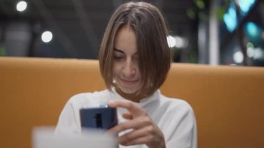 close up beautiful cheerful young woman sits at orange sofa in cafe and uses phone for chatting, texting, posting in social media. portrait of attractive caucasian female brunette with smartphone