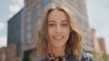 Slow motion close up portrait of attractive young woman in summer dress stands outdoors at city street, looking straight at camera and smiling. calm and confident person. wind blowing hair