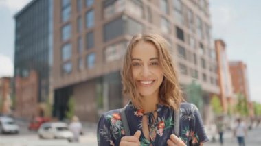 Slow motion close up portrait of attractive young woman in summer dress stands outdoors at city street, looking straight at camera and smiling. calm and confident person. wind blowing hair