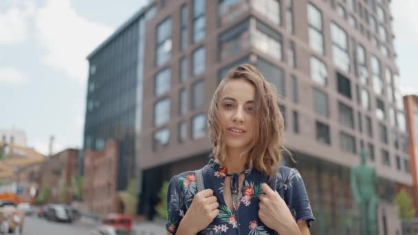 スローモーションの魅力的な若い女性は夏のドレスで街の通りで屋外に立って カメラをまっすぐ見ています 落ち着いて自信のある人 風吹いてる髪 — ストック動画