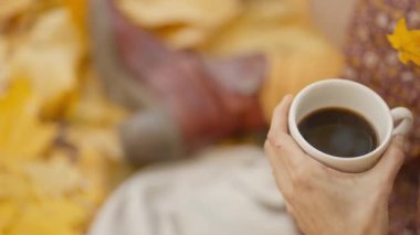 slow motion womans hands holds cup of coffee at picnic in bright autumn park with yellow foliage. stylish attractive girl sits on blanket in park at warm fall day and drinks coffee.