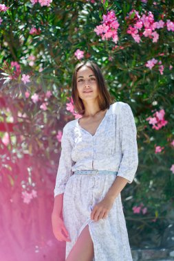 Spring time. Charming young lady wearing white dress posing near blooming pink flowers in garden. Concept of beauty and femininity