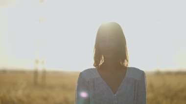 Outdoor portrait of charming happy woman smiling to camera, standing at wheat field in golden sun lights
