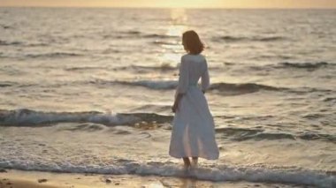 Woman in elegant dress standing at the sea beach during orange sundown. Nature and beauty concept. Girl silhouette at sunset