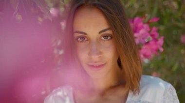 Yakından güzel bir portre. Siyah saçlı gülümseyen bir kız. Ağaçta duran pembe çiçeklere bakıyor..