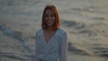 Zarif, uzun beyaz elbiseli bir kadın turuncu gün batımında deniz kıyısında yürüyor. Doğa ve güzellik kavramı. Gün batımında kız silueti