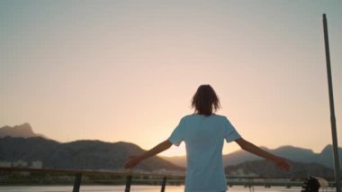 Happy carefree woman turning around to camera with hand, enjoying sunset time with orange sky. Sundown landscape with mountains and sea view