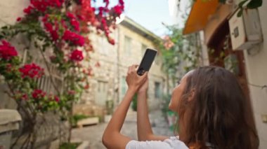 Ağır çekim. Elinde telefonla gezen çekici genç bir kadın fotoğrafçı cep telefonu kamerasında pembe ve kırmızı çiçeklerle güzel bir cephe fotoğrafı çekiyor..