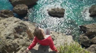 Türkiye 'nin Antalya kentindeki uçurumun kenarında oturan parlak kırmızı ceketli mutlu genç kadın gezginin portresi. Körfezdeki turkuaz Akdeniz manzarası