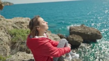 Türkiye 'nin Antalya kentindeki uçurumun kenarında oturan parlak kırmızı ceketli mutlu genç kadın gezginin portresi. Körfezdeki turkuaz Akdeniz manzarası