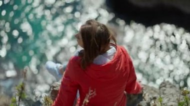 Türkiye 'nin Antalya kentindeki uçurumun kenarında oturan parlak kırmızı ceketli mutlu genç kadın gezginin portresi. Körfezdeki turkuaz Akdeniz manzarası