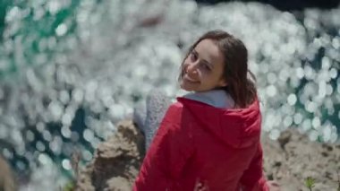 Türkiye 'nin Antalya kentindeki uçurumun kenarında oturan parlak kırmızı ceketli mutlu genç kadın gezginin portresi. Körfezdeki turkuaz Akdeniz manzarası