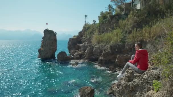 在土耳其安塔利亚 一位快乐快乐的年轻女性穿着鲜红色的夹克 坐在悬崖边 俯瞰大海 环湾绿松石地中海景致 — 图库视频影像
