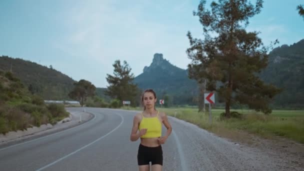 ロードを速く走る若いアスリートの女性のスローモーションの肖像画 ハードトレーニング レースの競争やマラソンの準備を取得 フィットガールで黒ショートパンツジョギングで夜明けに沿って緑のフィールド — ストック動画