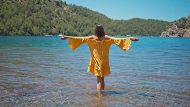 ビーチに立ち 海の上で晴れた日と太陽を楽しむオープン腕を持つ屈託のない女性オレンジのサンドレス 女の子はトルコの海沿岸を歩く — ストック動画