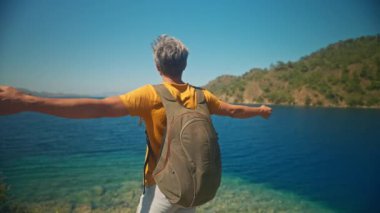 Sırt çantalı mutlu, orta yaşlı, gri saçlı gezgin turkuaz suyu ve cennet gibi boş deniz plajlı güzel deniz koyunun kıyısında uçurumun kenarında duruyor. Ağır çekim 4k, Türkiye 'de seyahat