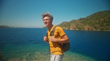 Sırt çantalı mutlu, orta yaşlı, gri saçlı gezgin turkuaz suyu ve cennet gibi boş deniz plajlı güzel deniz koyunun kıyısında uçurumun kenarında duruyor. Ağır çekim 4k, Türkiye 'de seyahat