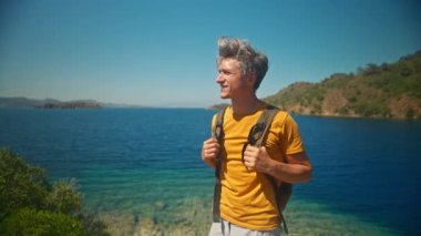 Sırt çantalı mutlu, orta yaşlı, gri saçlı gezgin turkuaz suyu ve cennet gibi boş deniz plajlı güzel deniz koyunun kıyısında uçurumun kenarında duruyor. Ağır çekim 4k, Türkiye 'de seyahat