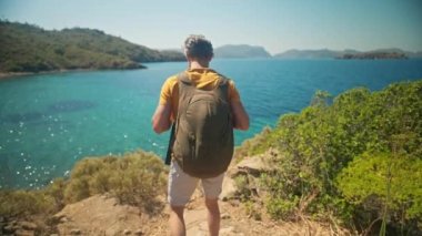 Sırt çantalı mutlu, orta yaşlı, gri saçlı gezgin turkuaz suyu ve cennet gibi boş deniz plajlı güzel deniz koyunun kıyısında uçurumun kenarında duruyor. Ağır çekim 4k, Türkiye 'de seyahat