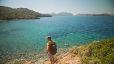 Sırt çantalı mutlu gezgin, temiz turkuaz suyu ve cennet gibi boş deniz plajları olan güzel deniz koyunun kıyısındaki uçurumda selfie ya da video çağrısı yapıyor. Ağır çekim, Türkiye 'de seyahat