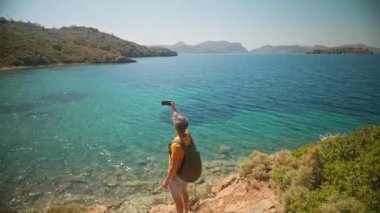 Sırt çantalı mutlu gezgin, temiz turkuaz suyu ve cennet gibi boş deniz plajları olan güzel deniz koyunun kıyısındaki uçurumda selfie ya da video çağrısı yapıyor. Ağır çekim, Türkiye 'de seyahat