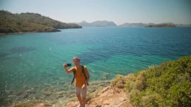 Sırt çantalı mutlu gezgin, temiz turkuaz suyu ve cennet gibi boş deniz plajları olan güzel deniz koyunun kıyısındaki uçurumda selfie ya da video çağrısı yapıyor. Ağır çekim, Türkiye 'de seyahat