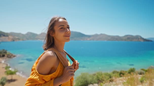 オレンジ色のサンドレスの魅力的なスリムな日焼けした女性旅行者はターコイズブルーの水と楽園の空の海のビーチと美しい湾の海岸に立っています 女の子はトルコの海沿岸 マルマリス地方を歩く — ストック動画
