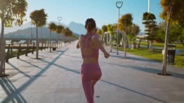 Arkaya bakan yürüyüş kamerası, gün batımında gezinti güvertesinde koşan genç fitness sporcusunu çekti.